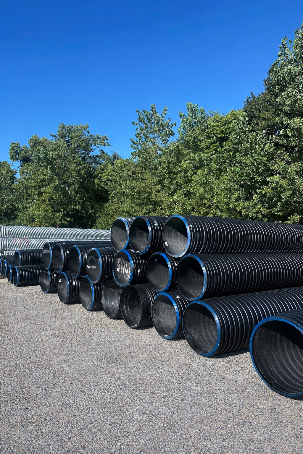 Stacks of large black corrugated pipes outdoors on gravel under a clear blue sky.