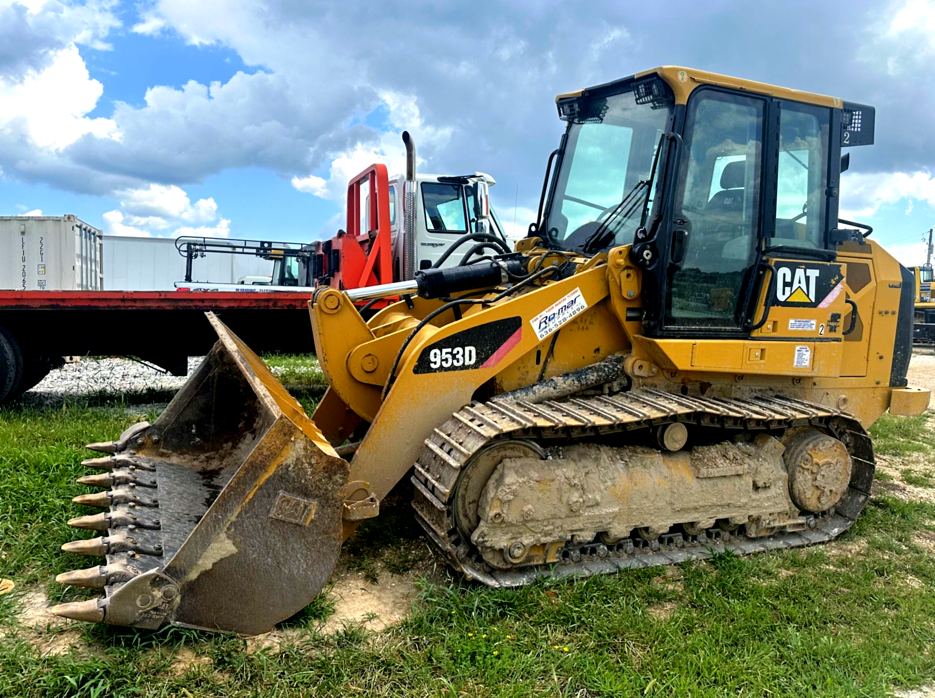 Yellow CAT track loader with bucket and grapple attachment on grass near a trailer under a cloudy sky