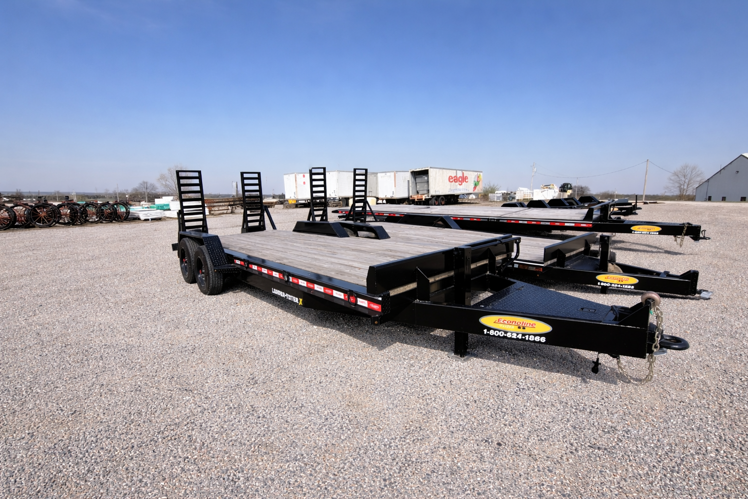Two black flatbed utility trailers parked on a gravel lot under a clear blue sky.