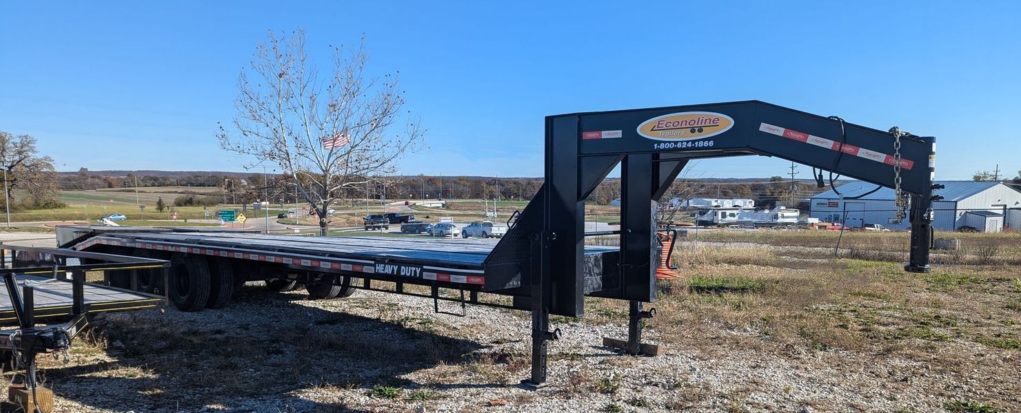 Black gooseneck flatbed trailer on a gravel lot with a blue sky background.