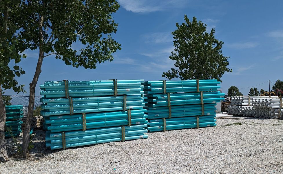 Stacks of teal pipes on a gravel lot, under a blue sky with trees.