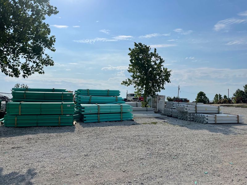Piles of green and gray lumber on a gravel lot under a blue sky with trees.