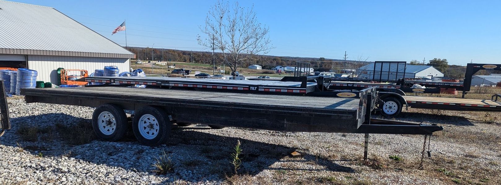 A long, flatbed trailer sits on a gravel lot in front of a blue sky.