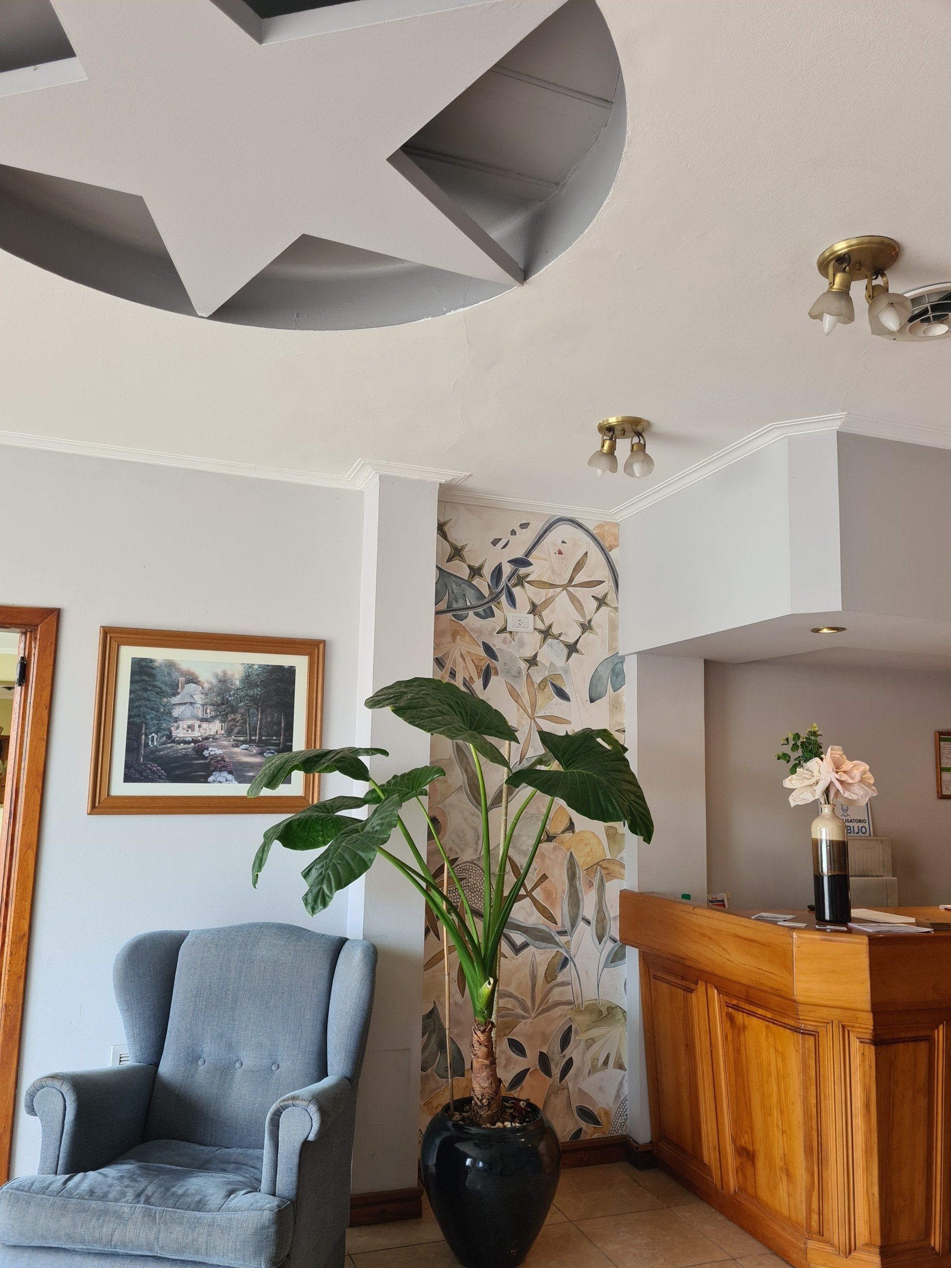 Vestíbulo con un sillón gris, una maceta grande y un mostrador de recepción. Un cuadro cuelga de la pared.