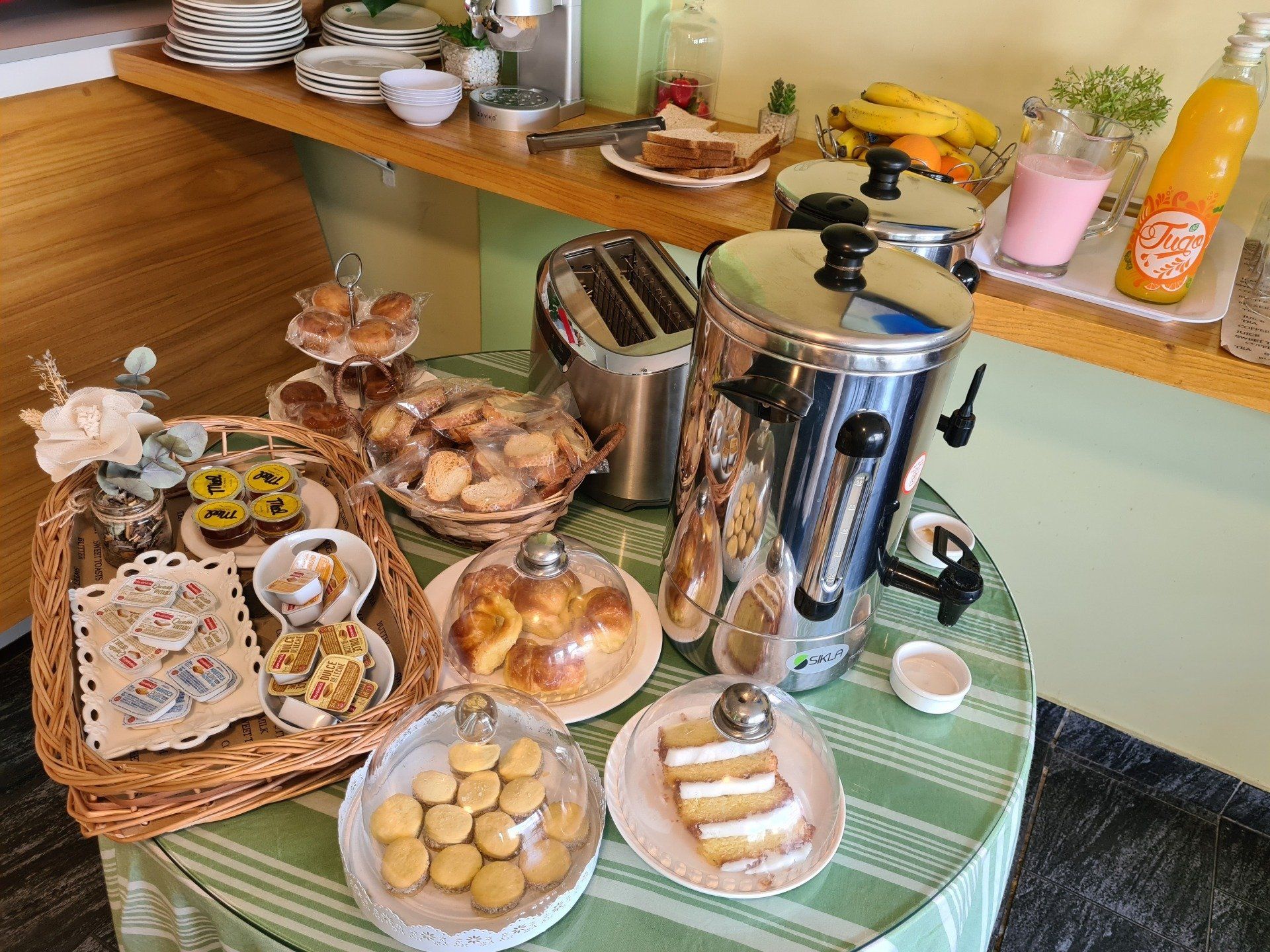 Desayuno buffet con bollería, bebidas y cafetera en mesa redonda, ambiente luminoso y acogedor.