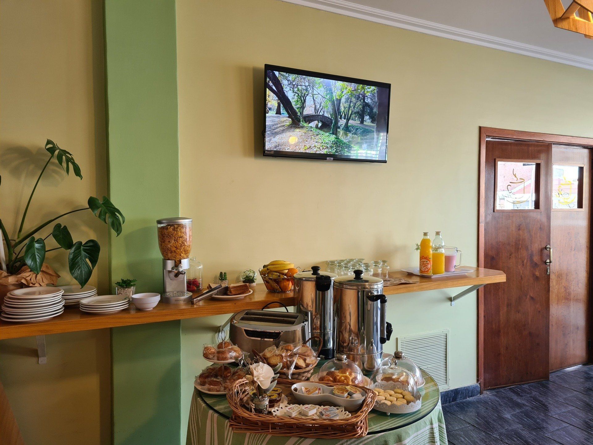 Desayuno buffet con cereales, fruta, pan, zumo y café en una mesa y estanterías.