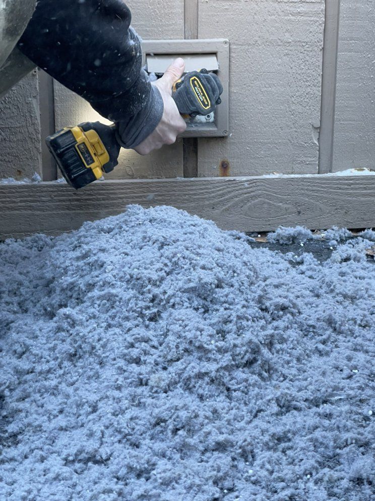 Person using a drill on a vent cover next to a pile of cellulose insulation on a wooden surface.