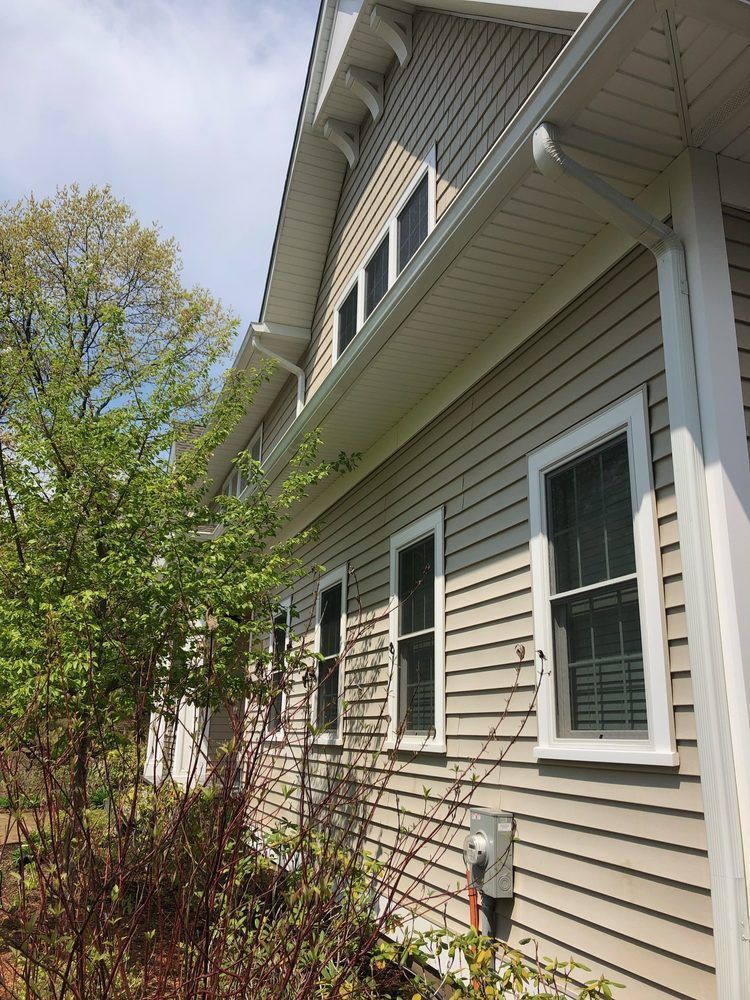 Beige house exterior with windows, siding, and gutters, set against a partly sunny sky, foliage in the foreground.