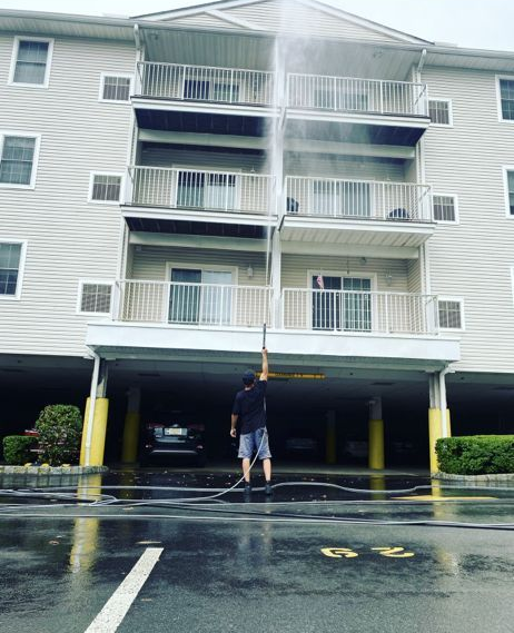 Person pressure washing the exterior of a three-story beige apartment building with balconies. Wet ground.