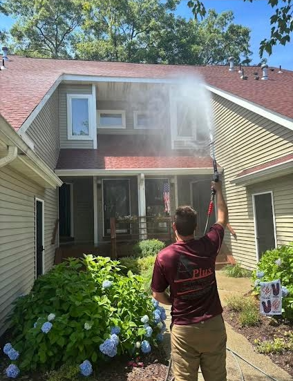 Man power washing a two-story beige house with a red roof, spraying water towards the front. Blue hydrangeas are in foreground.