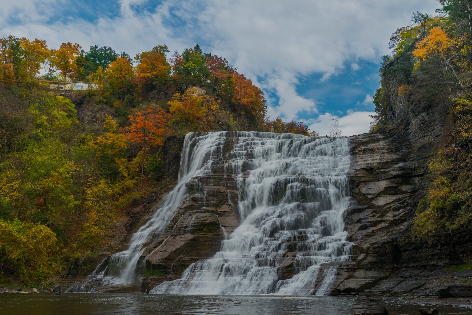 Wide waterfall cascading over rocky cliffs with autumn-colored trees and blue sky above