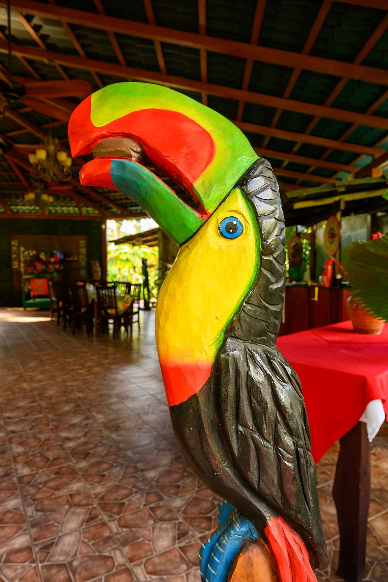 Colorful carved toucan statue in a restaurant with a red tablecloth and tiled floor.