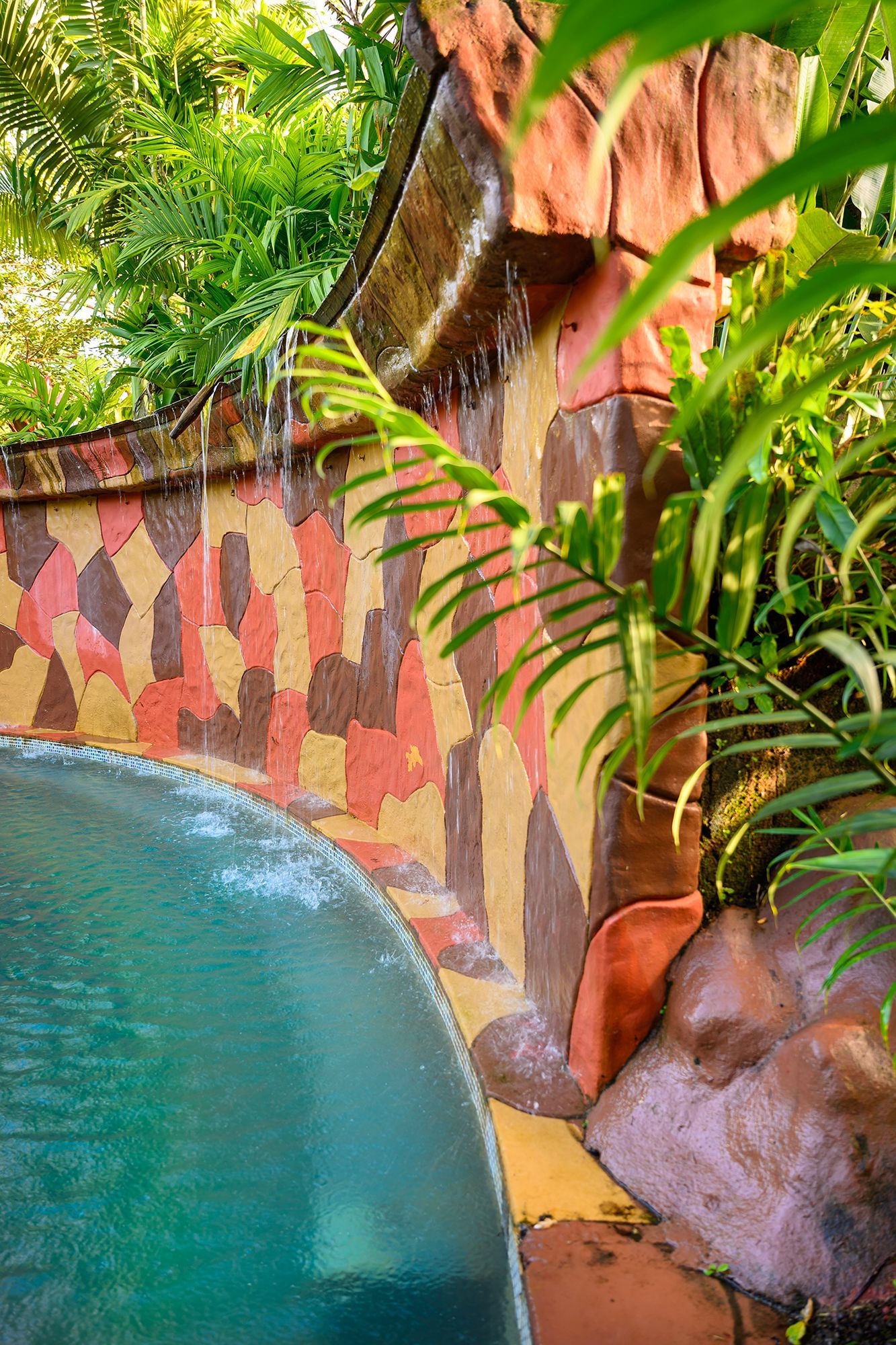 Waterfall cascading into a pool with a colorful patterned wall, surrounded by lush green foliage.