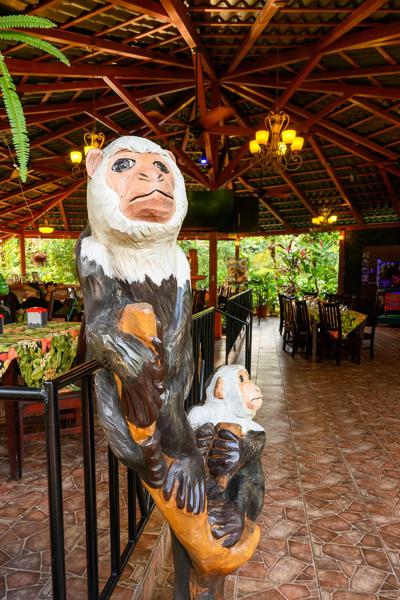 Statue of a monkey and child in an open-air restaurant. Tables, chairs, and greenery in the background.