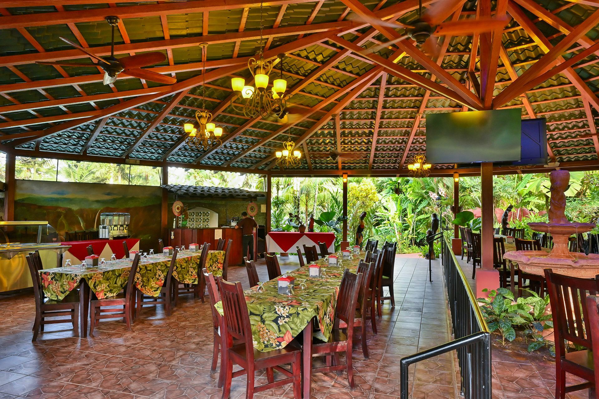 Open-air restaurant with tables set for dining under a thatched roof. Green foliage visible outside.