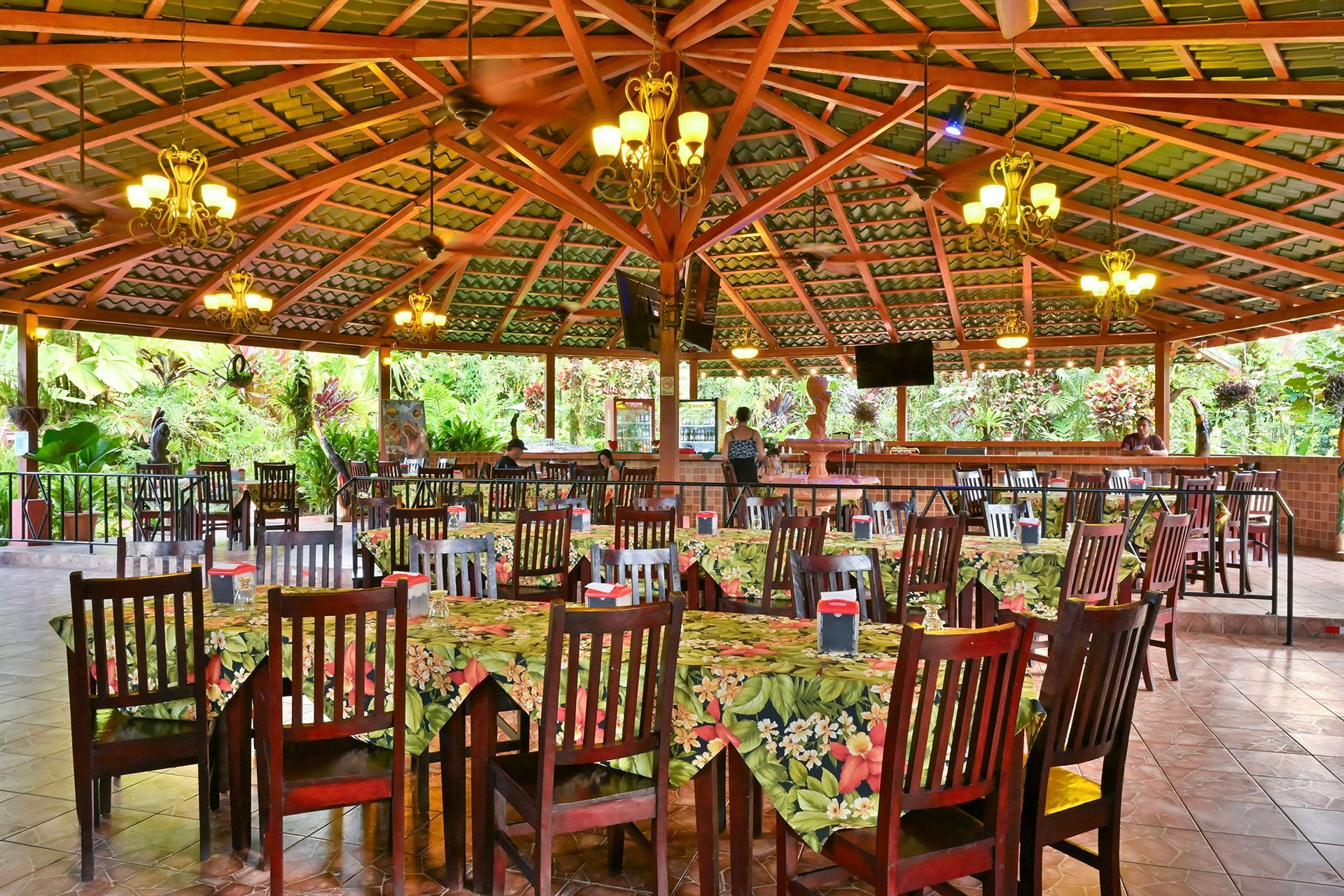 Restaurant with tables and chairs under a thatched roof with hanging lights; a bar and television visible.
