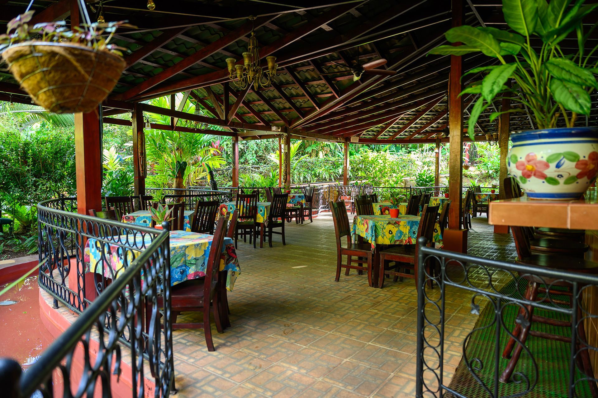 Outdoor restaurant with tables set under a wooden roof. Lush green plants surround the open-air seating area.