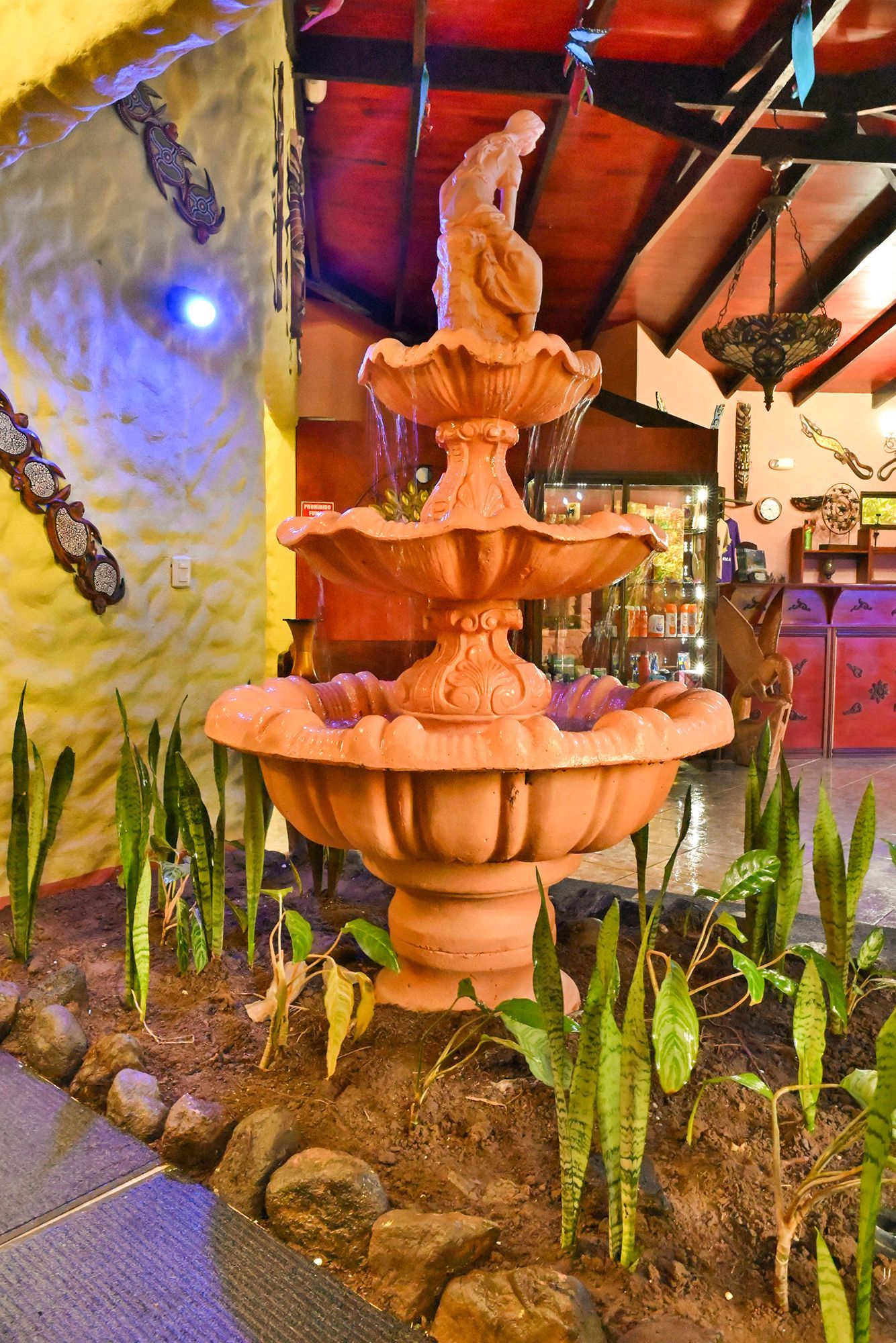 Terracotta fountain with a statue, surrounded by plants, indoors.