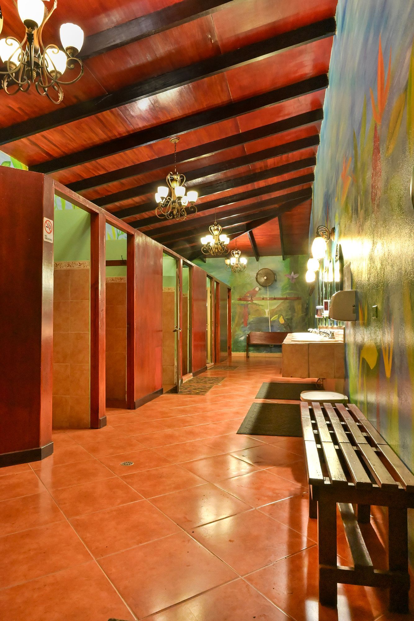 Public restroom with red tiled floor, stalls, wood bench, and ornate chandeliers.