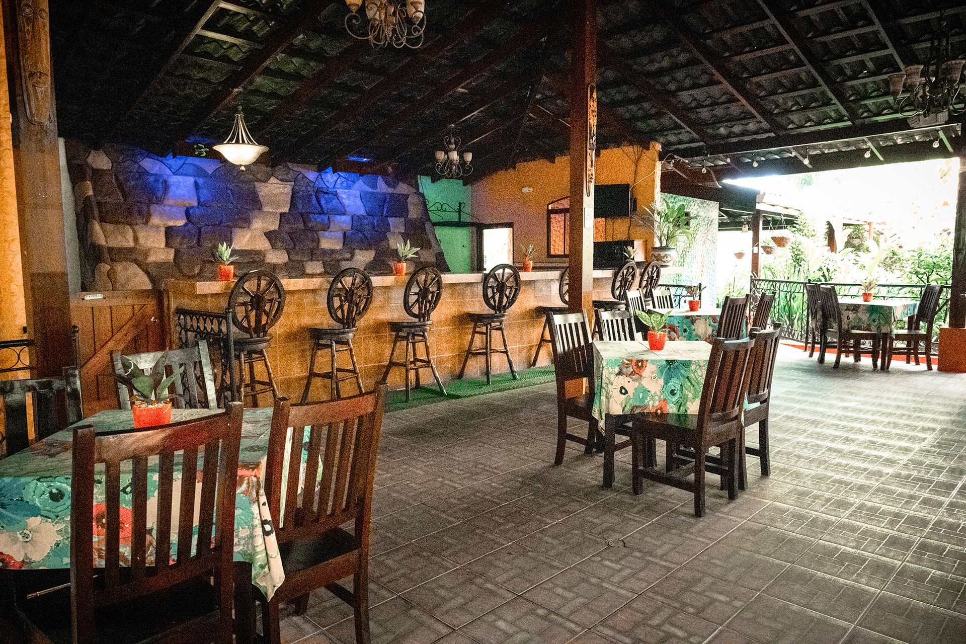 Restaurant interior with bar and tables. Wood chairs and tables, patterned tablecloths, open-air setting.