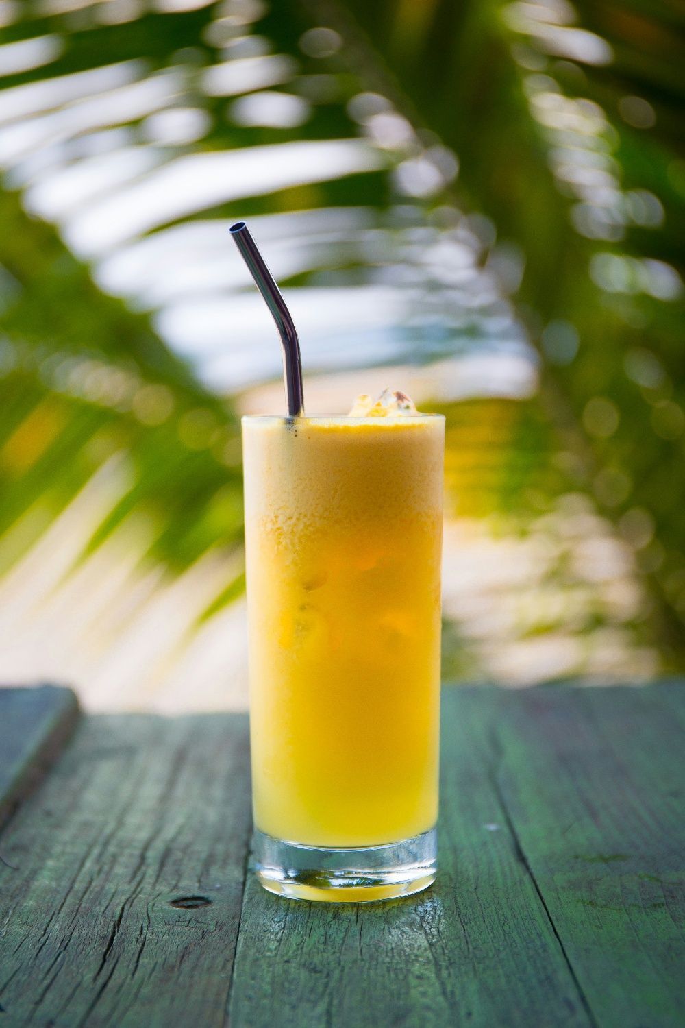 Tall glass of yellow drink with ice, straw, on a wooden table, blurry tropical background.