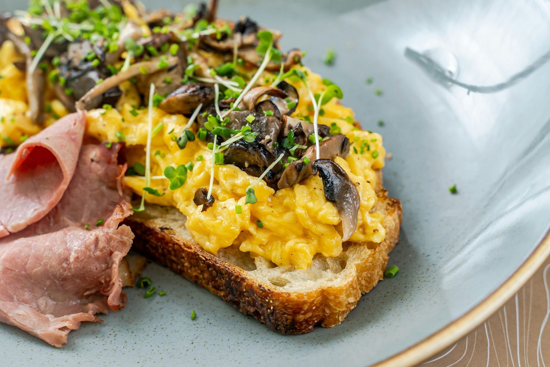 Scrambled eggs with mushrooms and microgreens on toast, with ham on a blue plate.