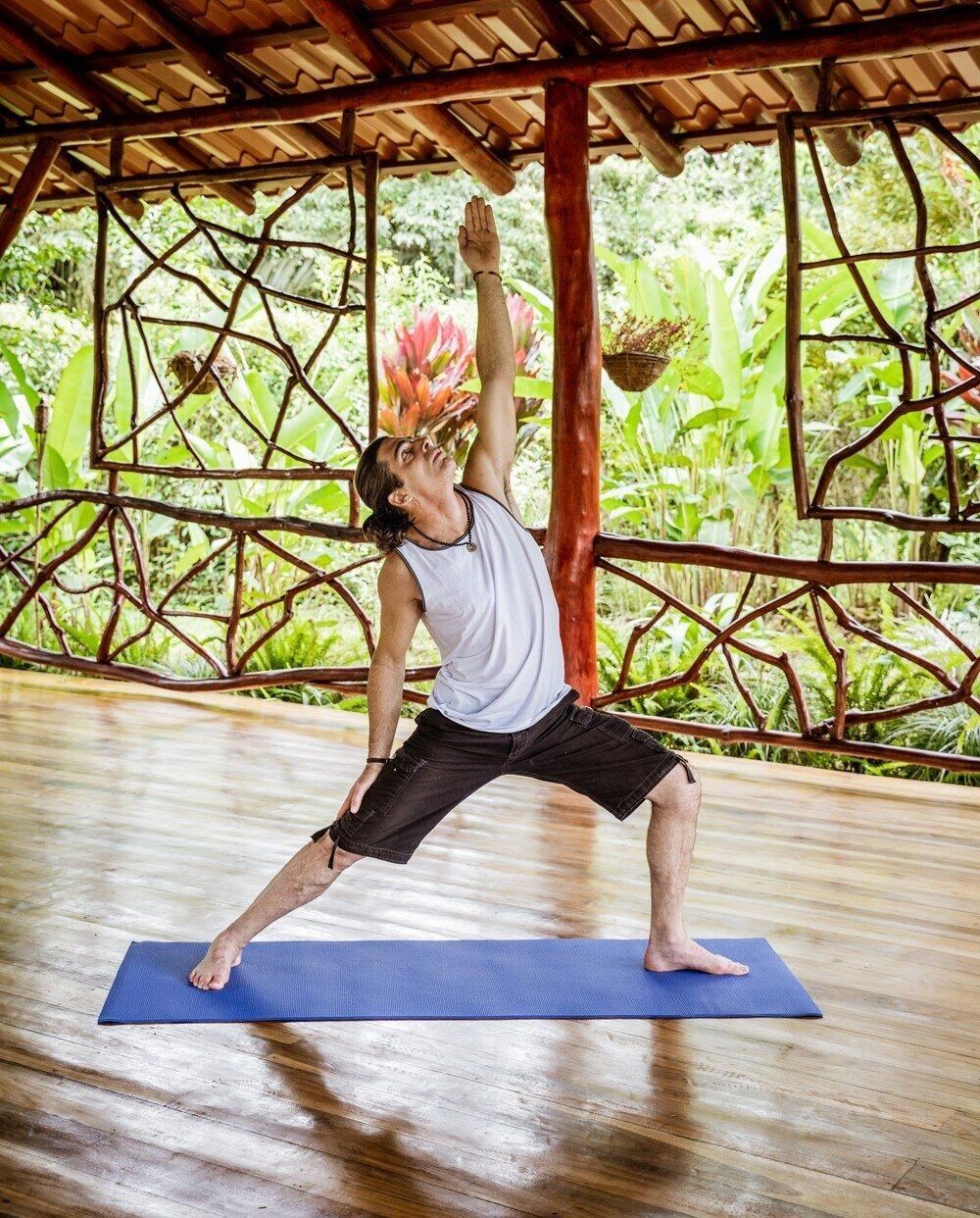 Man in yoga pose on mat, arms raised, outdoor wooden setting.