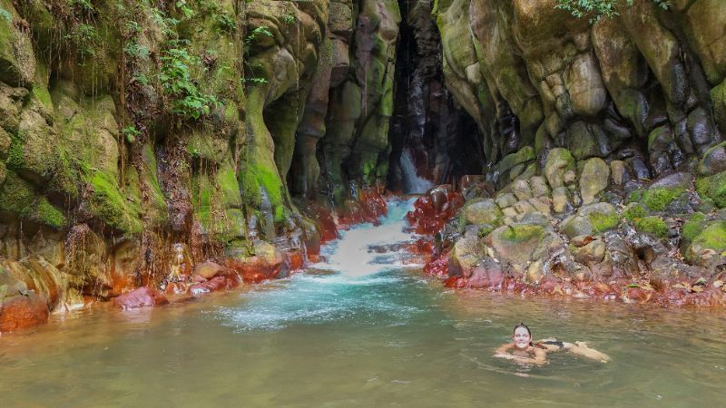 Waterfall cascading into a turquoise pool, surrounded by mossy rocks. Person swimming.