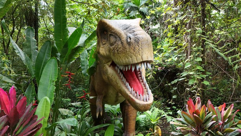 Dinosaur statue with open mouth in a lush green jungle setting, with red and pink foliage.