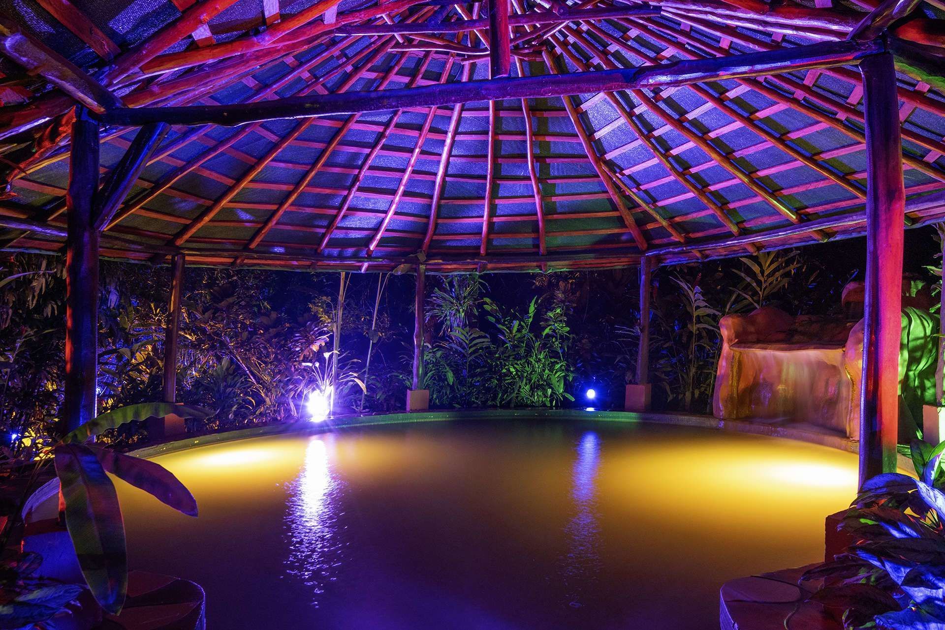 Illuminated gazebo over a pool of water at night, glowing with blue and yellow lights.