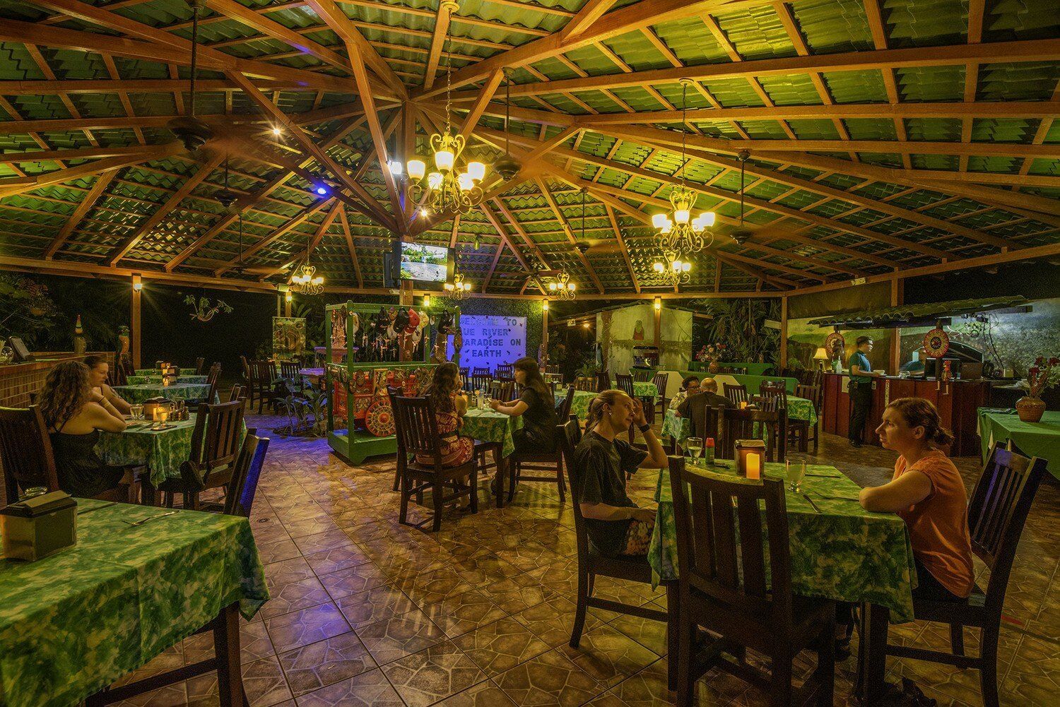 Indoor restaurant with a thatched roof, tables with green tablecloths, and people dining under soft lighting.