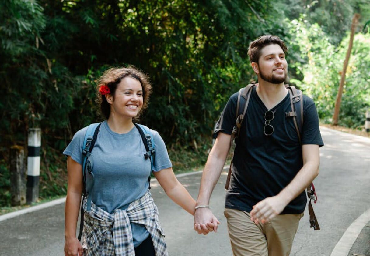 Couple walking hand in hand, smiling, on a path through a wooded area. They wear backpacks.