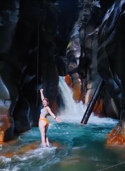 Woman in swimsuit stands in a turquoise river, arms outstretched. Waterfall in a dark, rocky gorge.