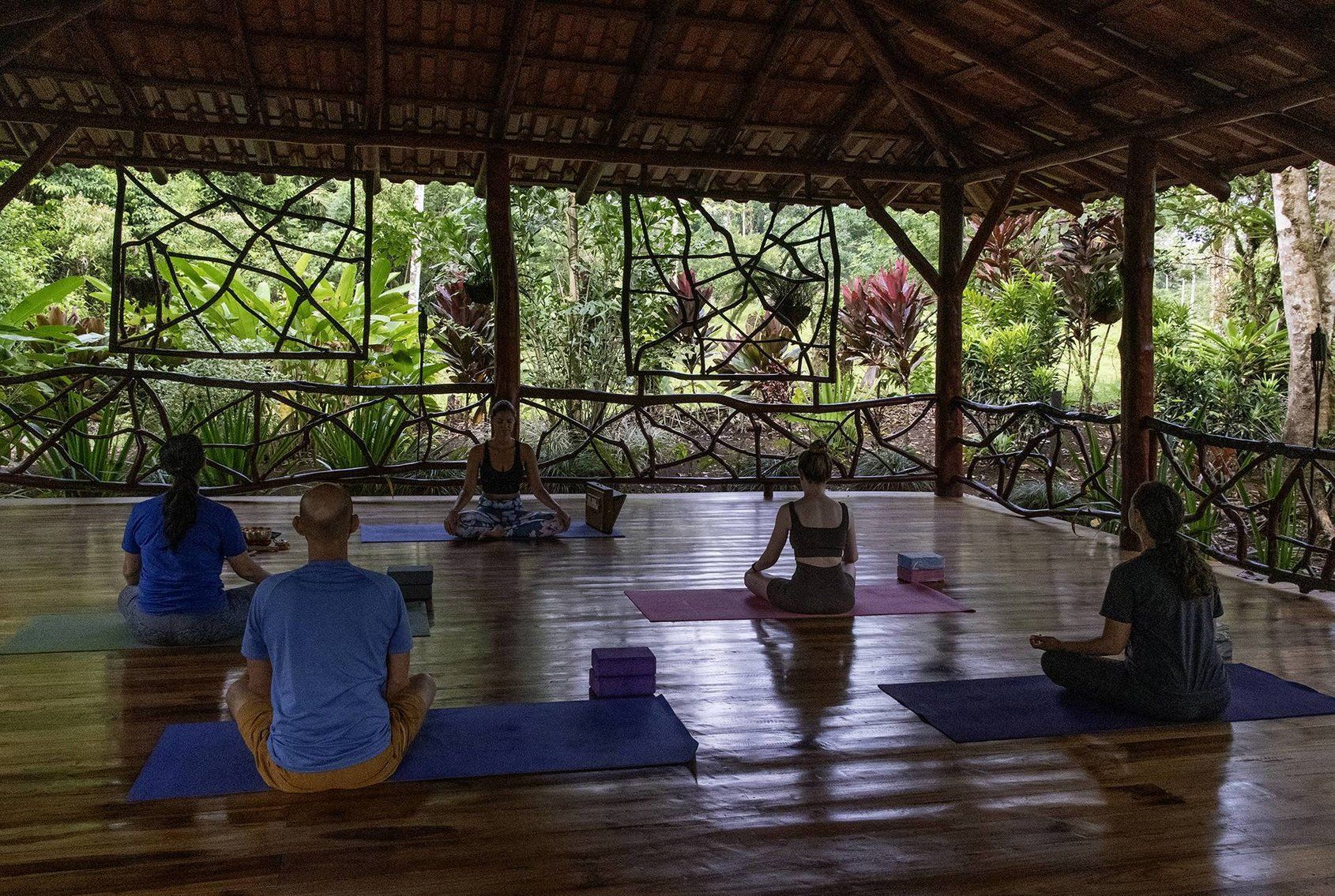 People in yoga poses on mats under a wooden structure; nature in background.