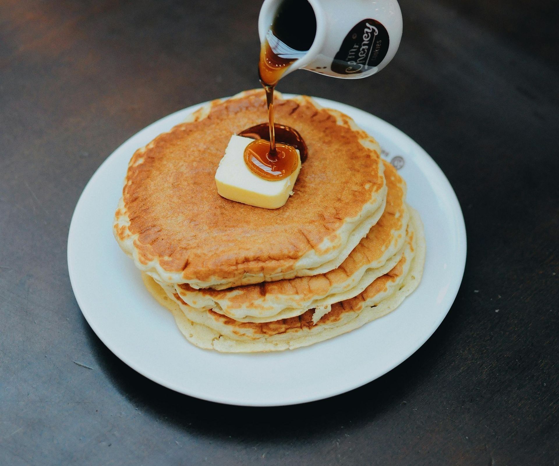 Stack of golden pancakes with melting butter, being drizzled with syrup from a white pitcher.