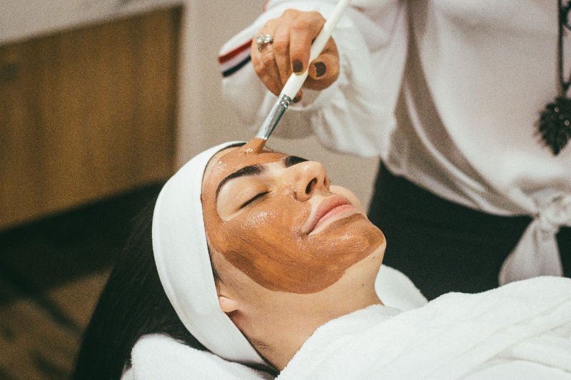 Person receiving a facial mask application with a brush in a spa-like setting.