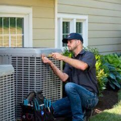 A man is kneeling down in front of a house fixing an air conditioner.