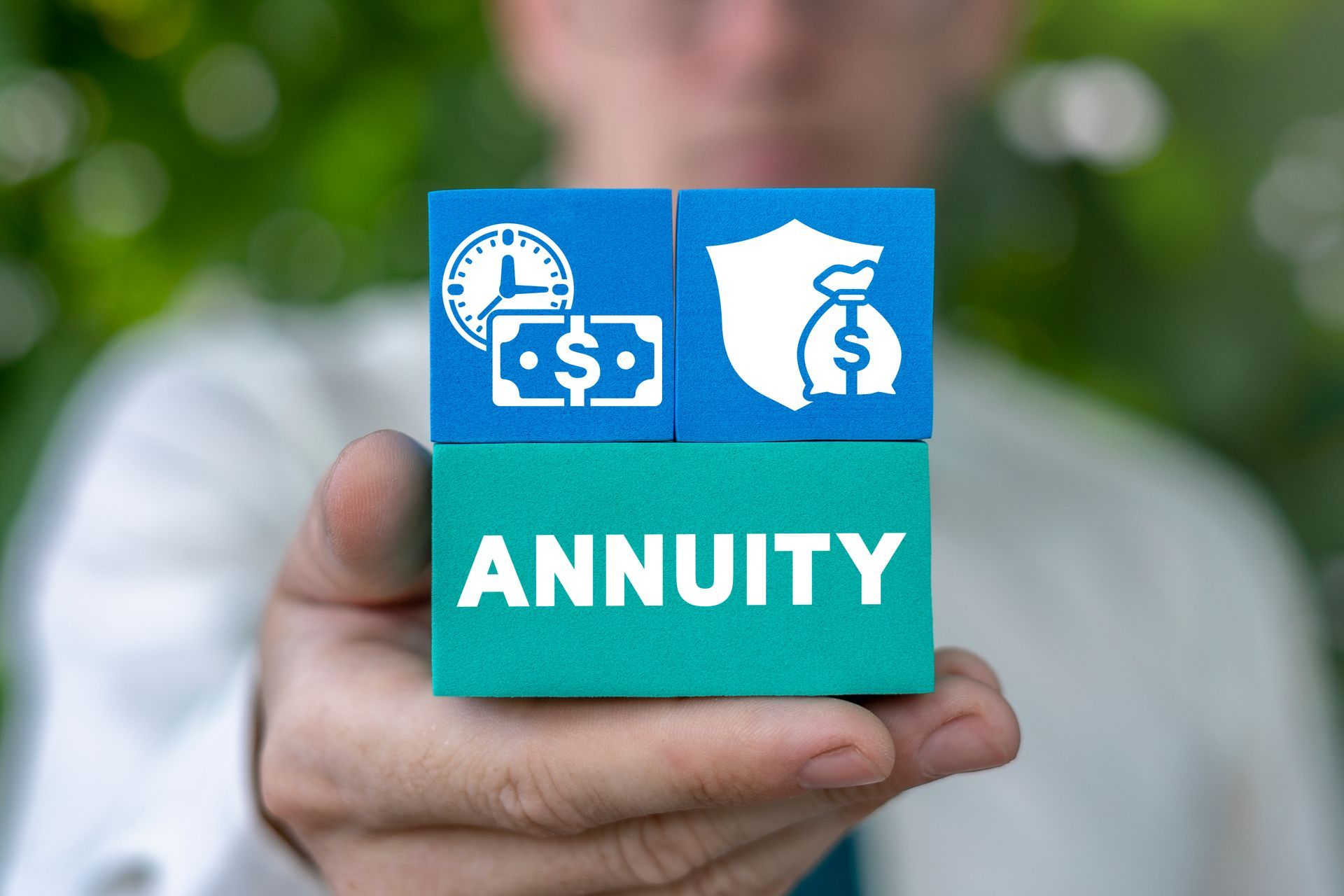 Hand holding a blue block labeled “ANNUITY” with money and savings icons against a blurred background