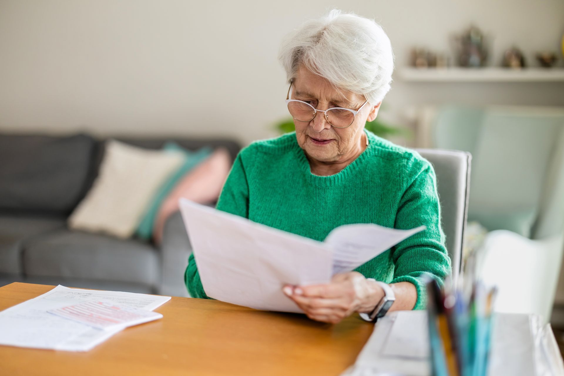 Elderly woman in a green sweater reading papers at a table in a bright living room