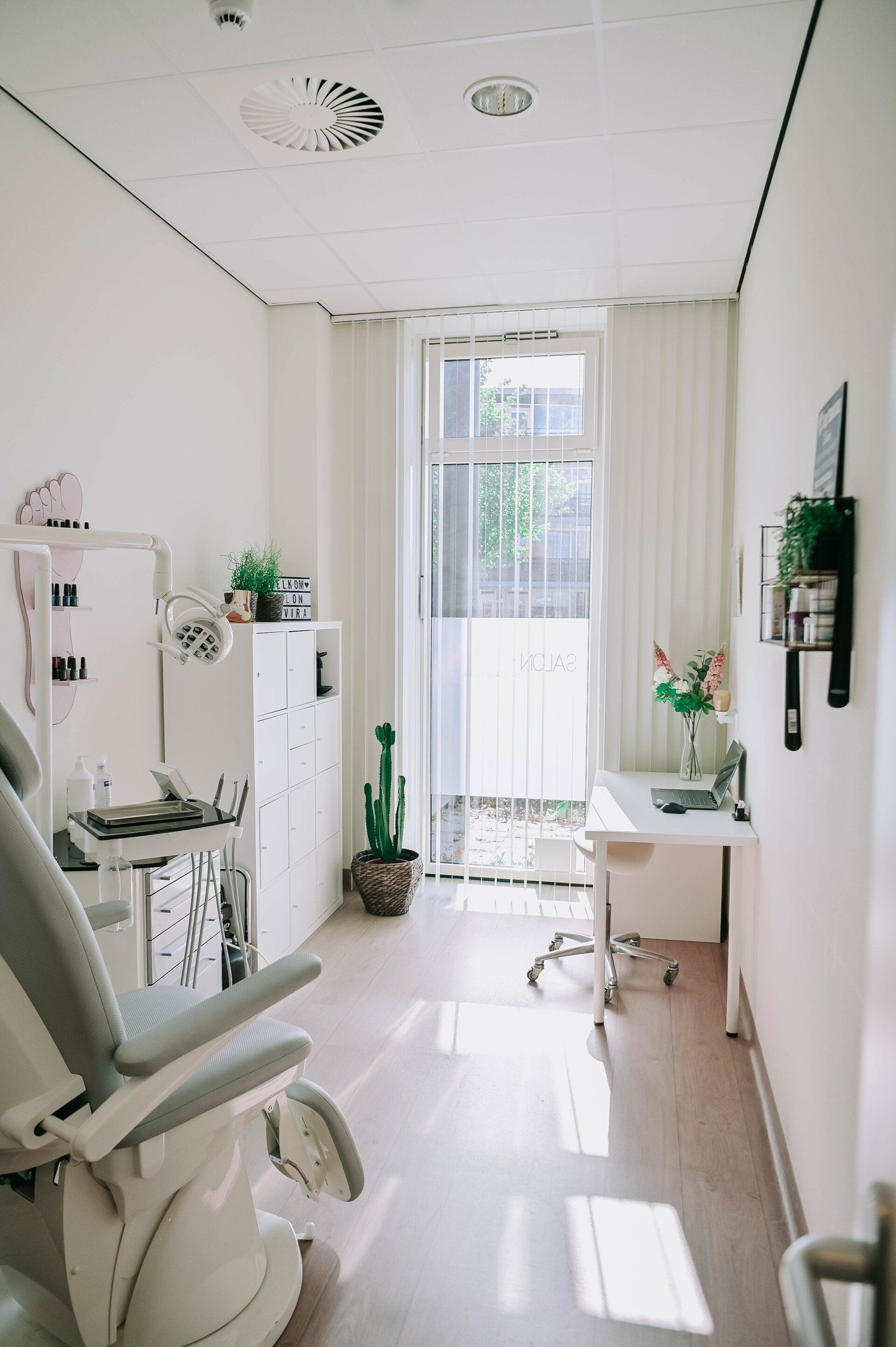 Lichte, witte behandelkamer met tandartsstoel, bureau en groot raam. Zonlicht stroomt naar binnen.