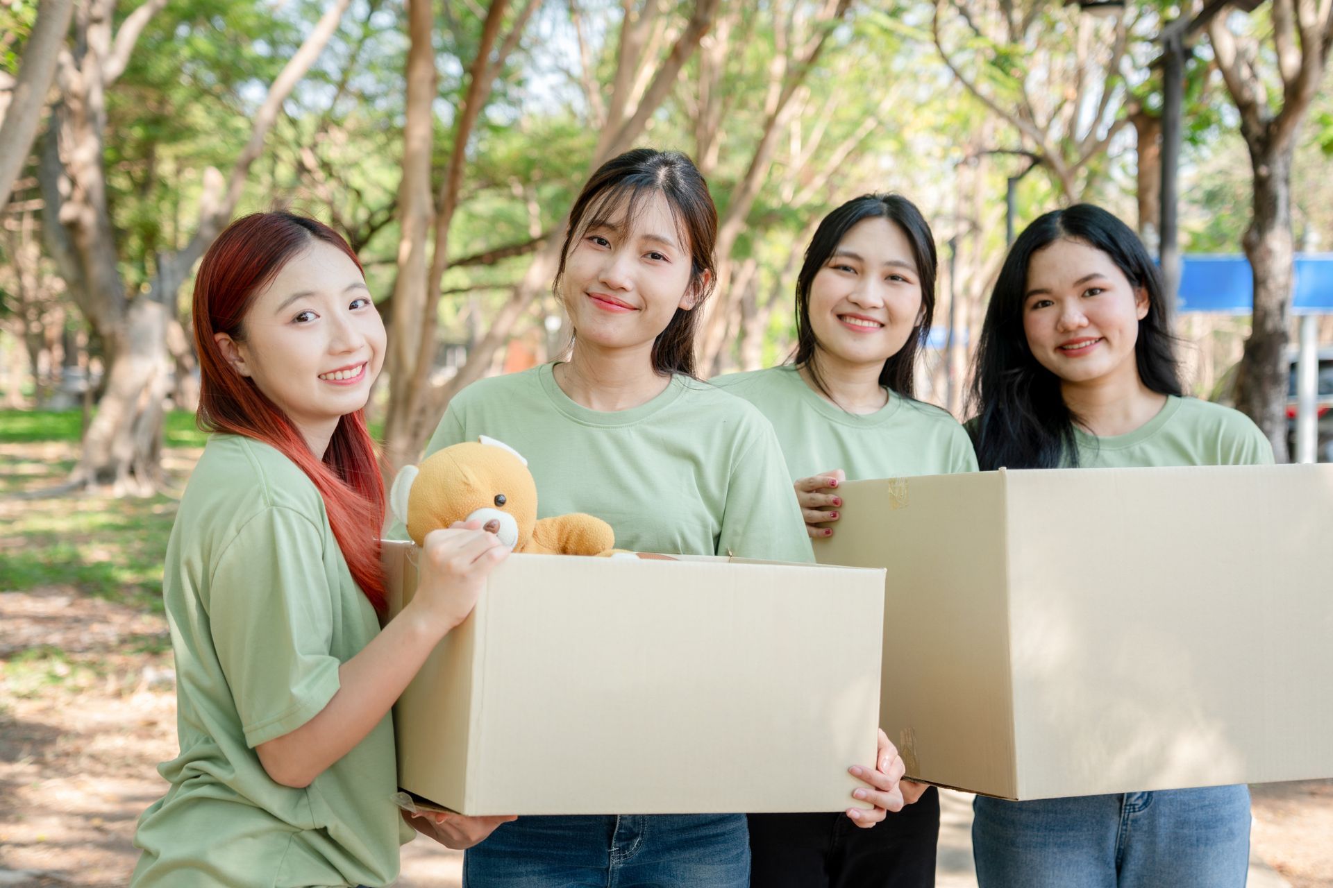 Four young women in green shirts smiling, holding cardboard boxes outdoors.