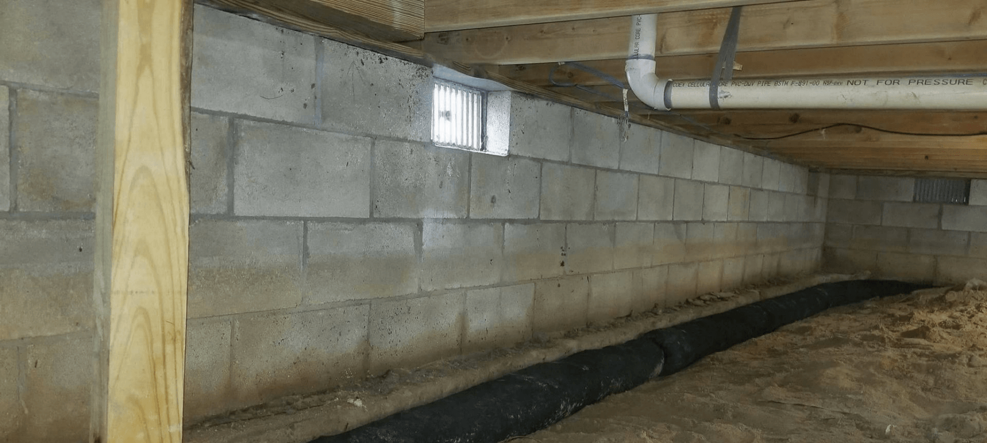Crawlspace under a house with cinder block walls, wooden beams, and a small window opening