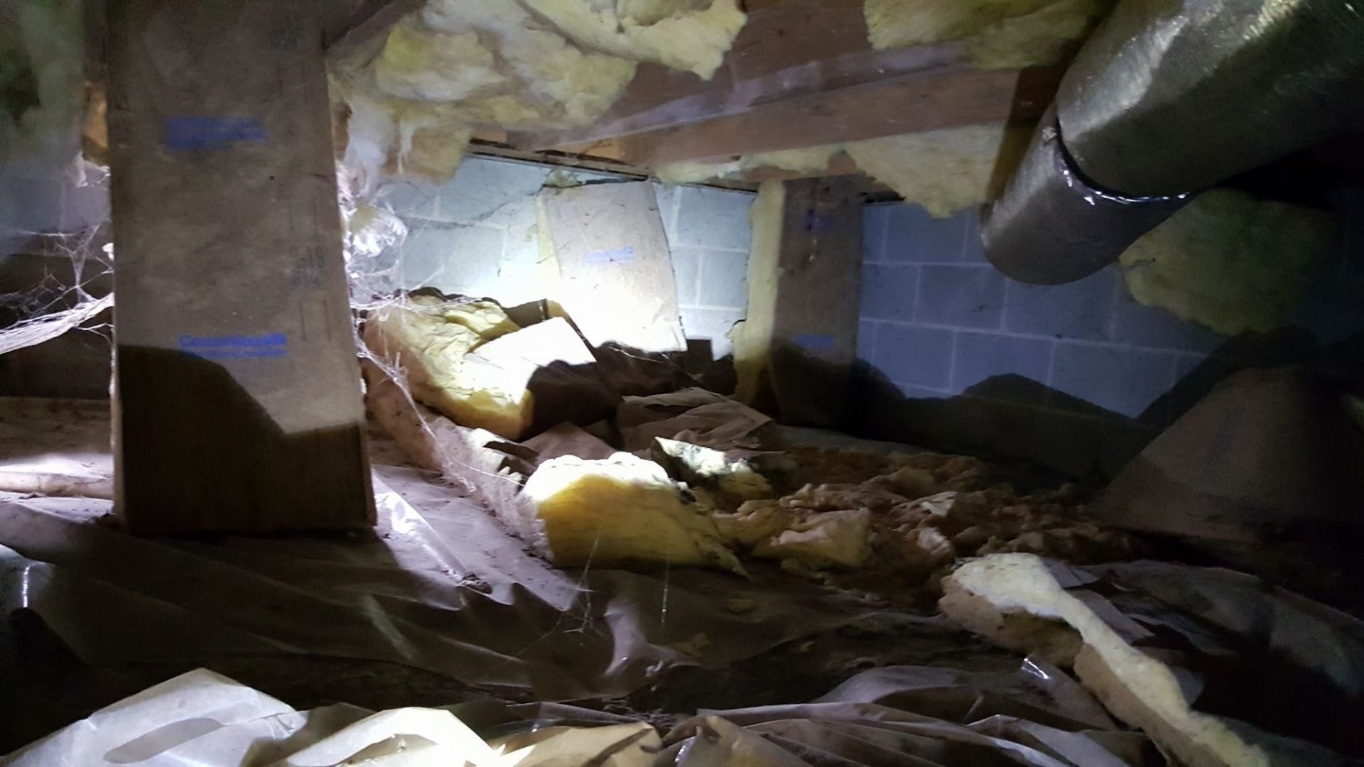 Collapsed attic with insulation debris, exposed beams, and a large pipe in a dimly lit crawlspace
