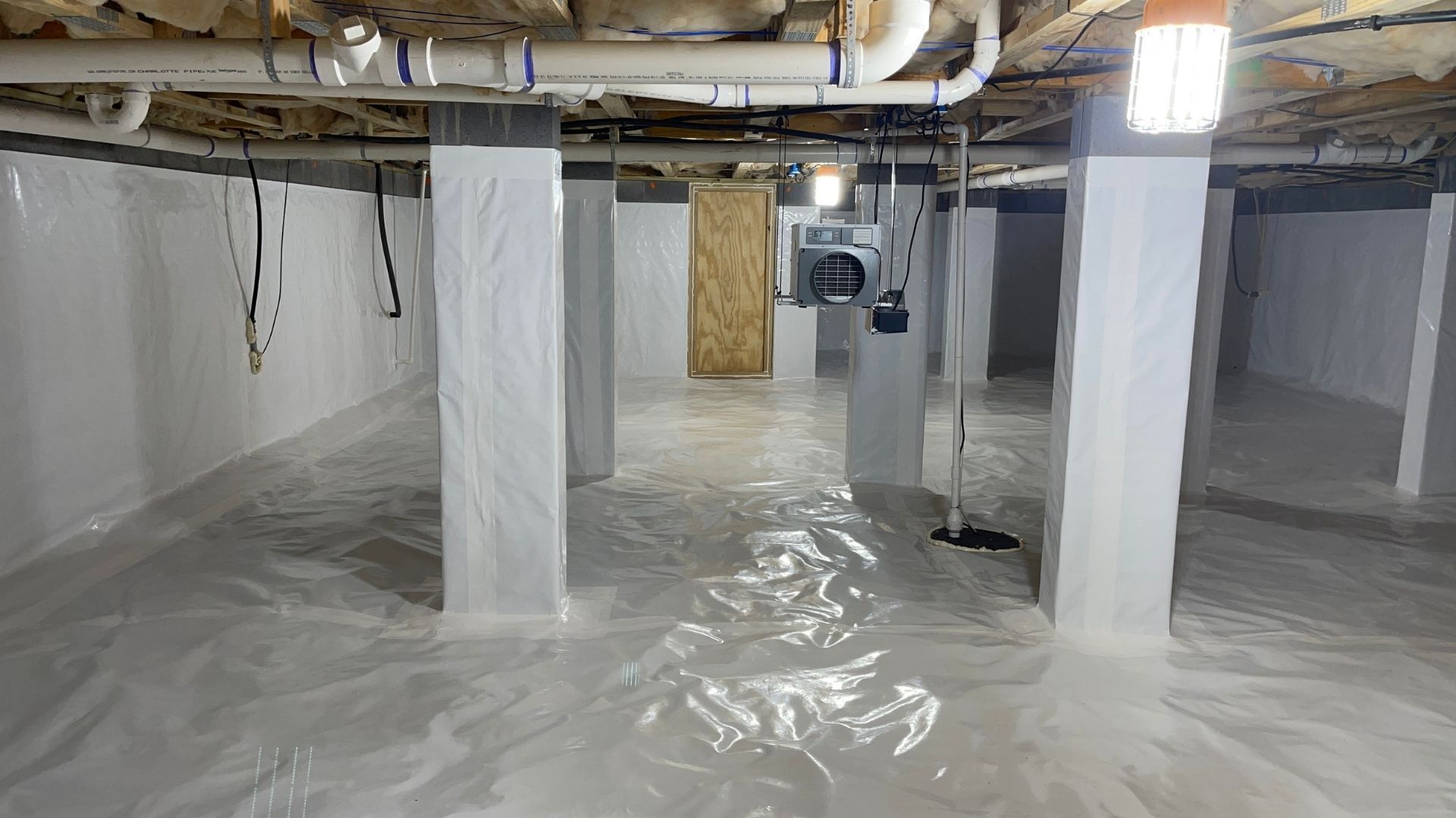 Basement crawlspace lined with white plastic sheeting, exposed pipes, and support columns