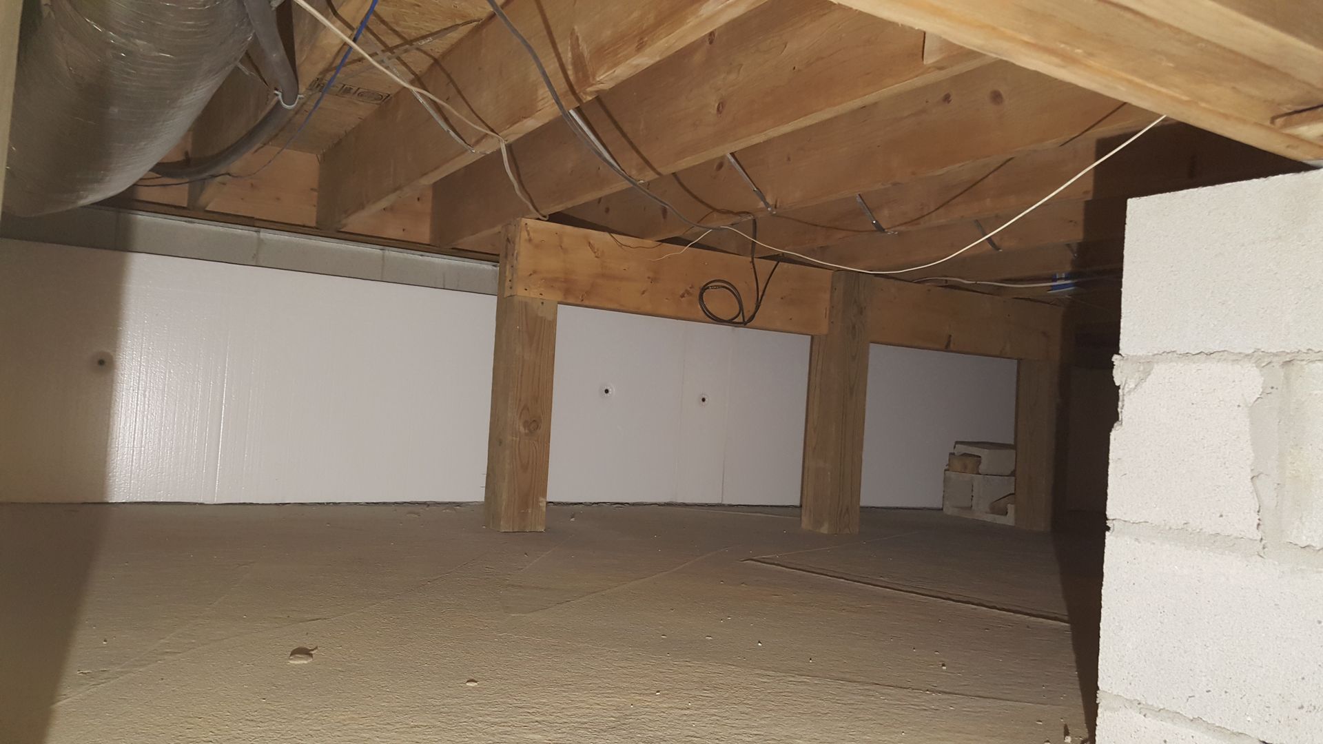 Attic crawlspace with exposed wooden beams, white wall panels, and unfinished flooring