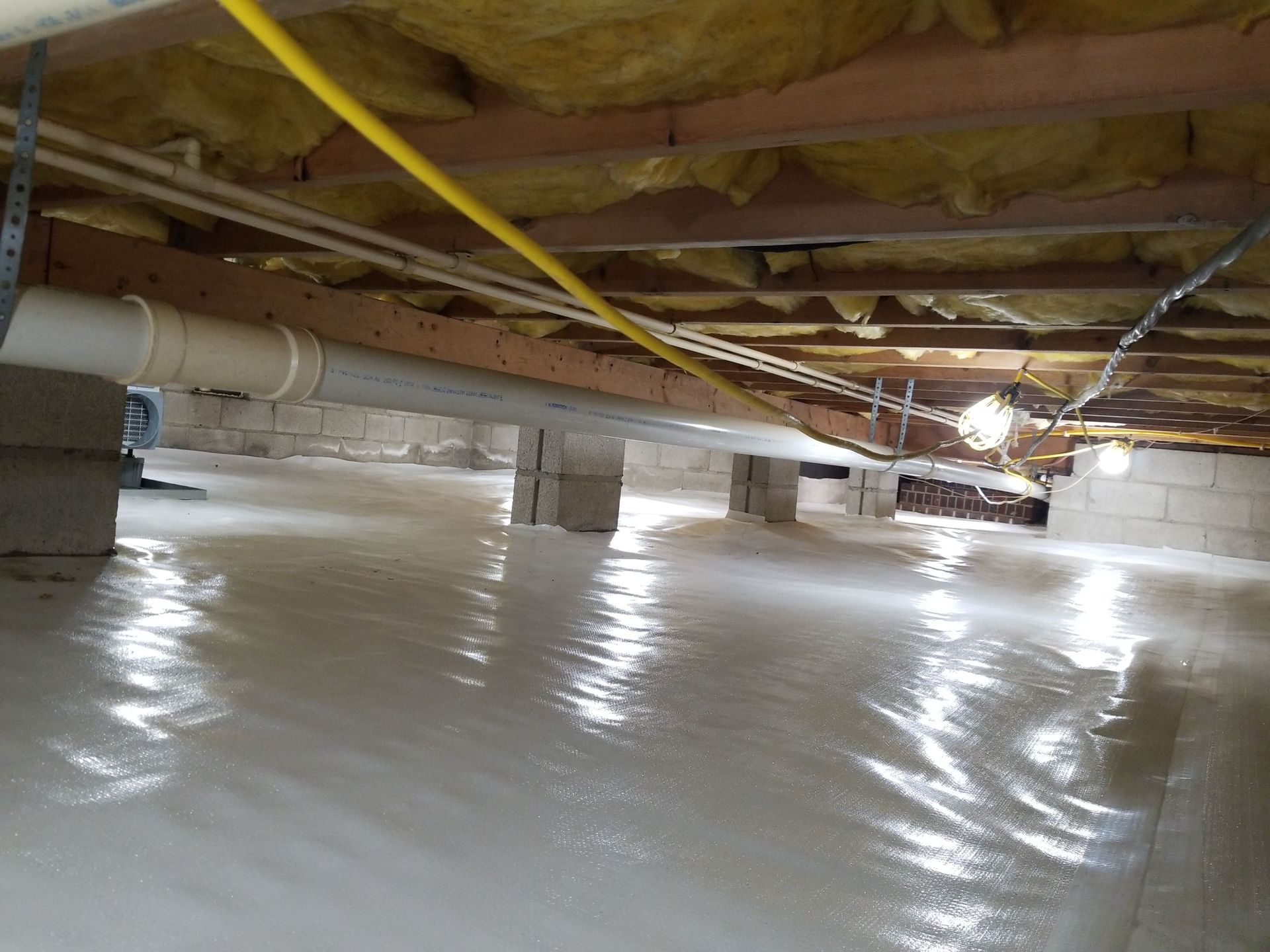Low crawl space with white vapor barrier, exposed wooden beams, and yellow utility lines under a house