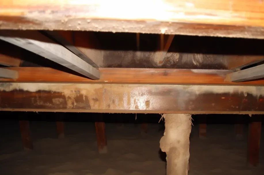Inside a crawlspace, rusty metal beams and a wooden floor frame a central support post.