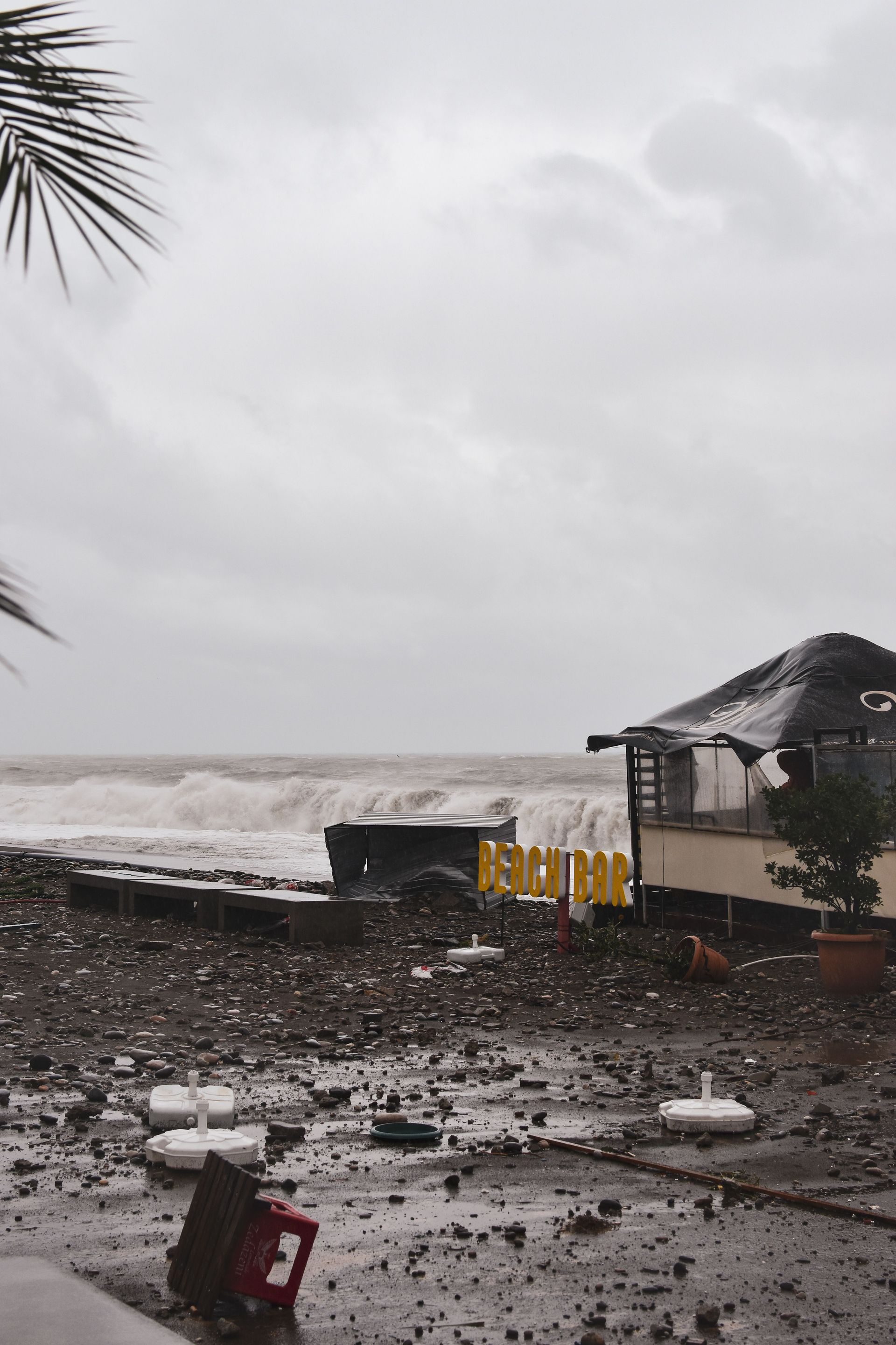 Storm-battered beach with waves, debris, and a damaged seaside hut under a gray sky