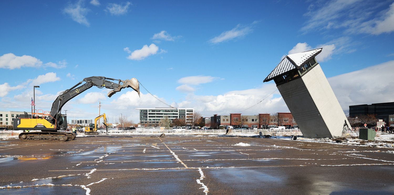 Construction excavator demolishing a leaning white tower in a snowy lot under a blue sky