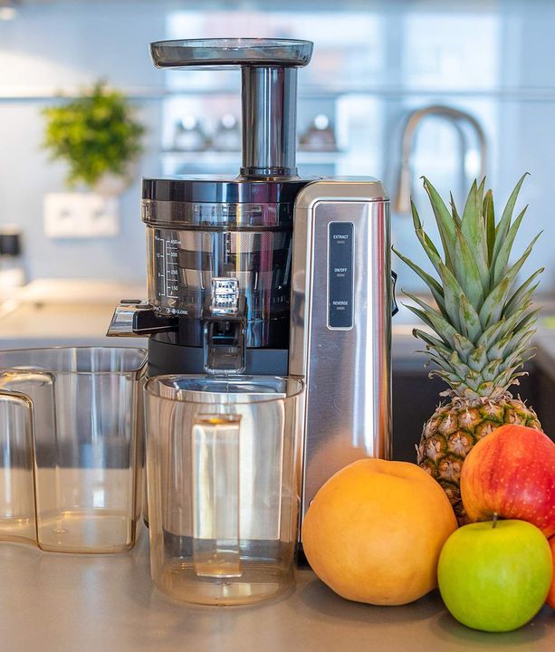 Juicer on kitchen counter with glasses, pineapple, oranges, and an apple